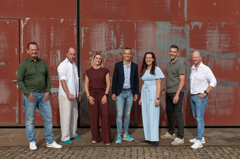 De zeven bestuursleden van Stichting Baggerfestival, naast elkaar voor een roestbruine loodsdeur